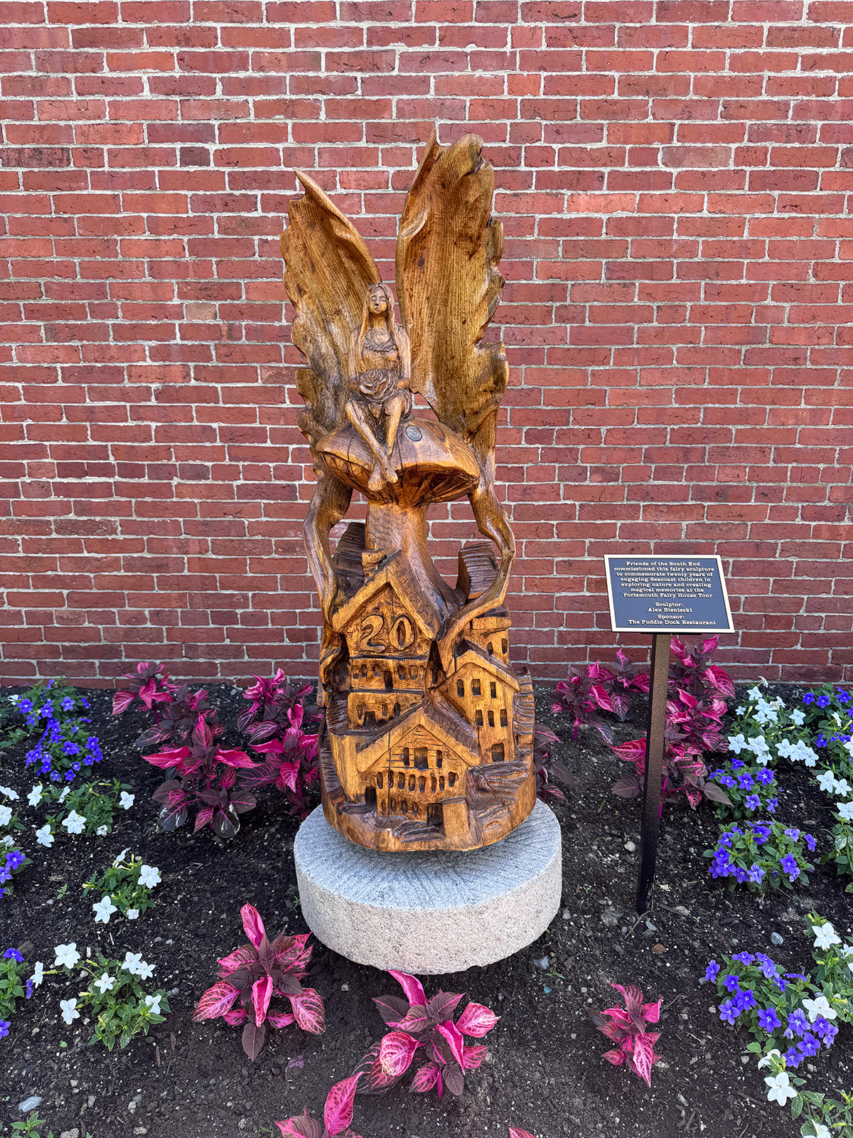 an image of the carved wooden fairy house sculpture which depicts a fairy with wings spread wide sitting on a mushroom atop a cluster of houses, one with the big number 20 carved into it.