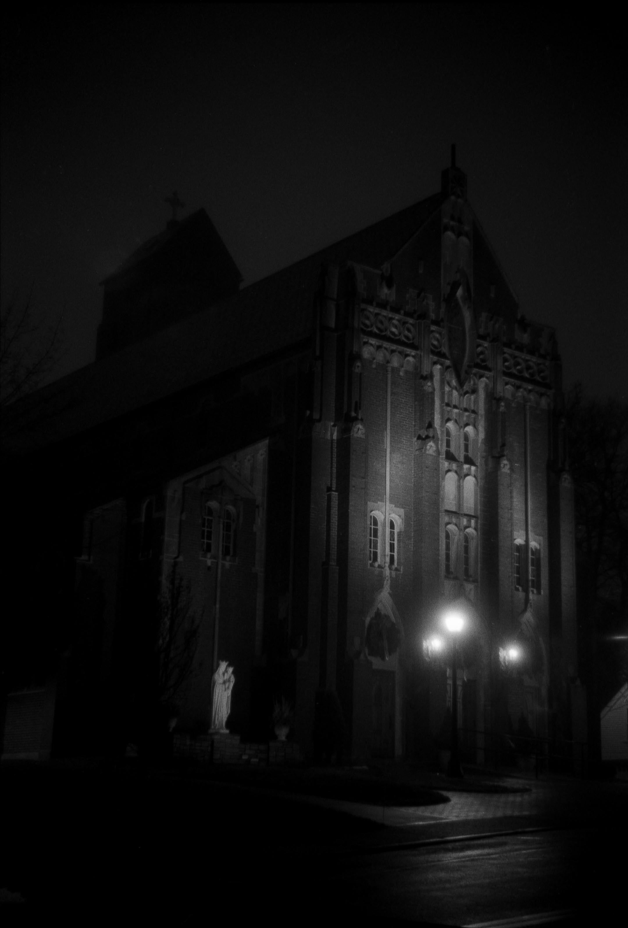 A gothic church at night in black and white illuminated only by street lamps with a lone statue in the near distance.