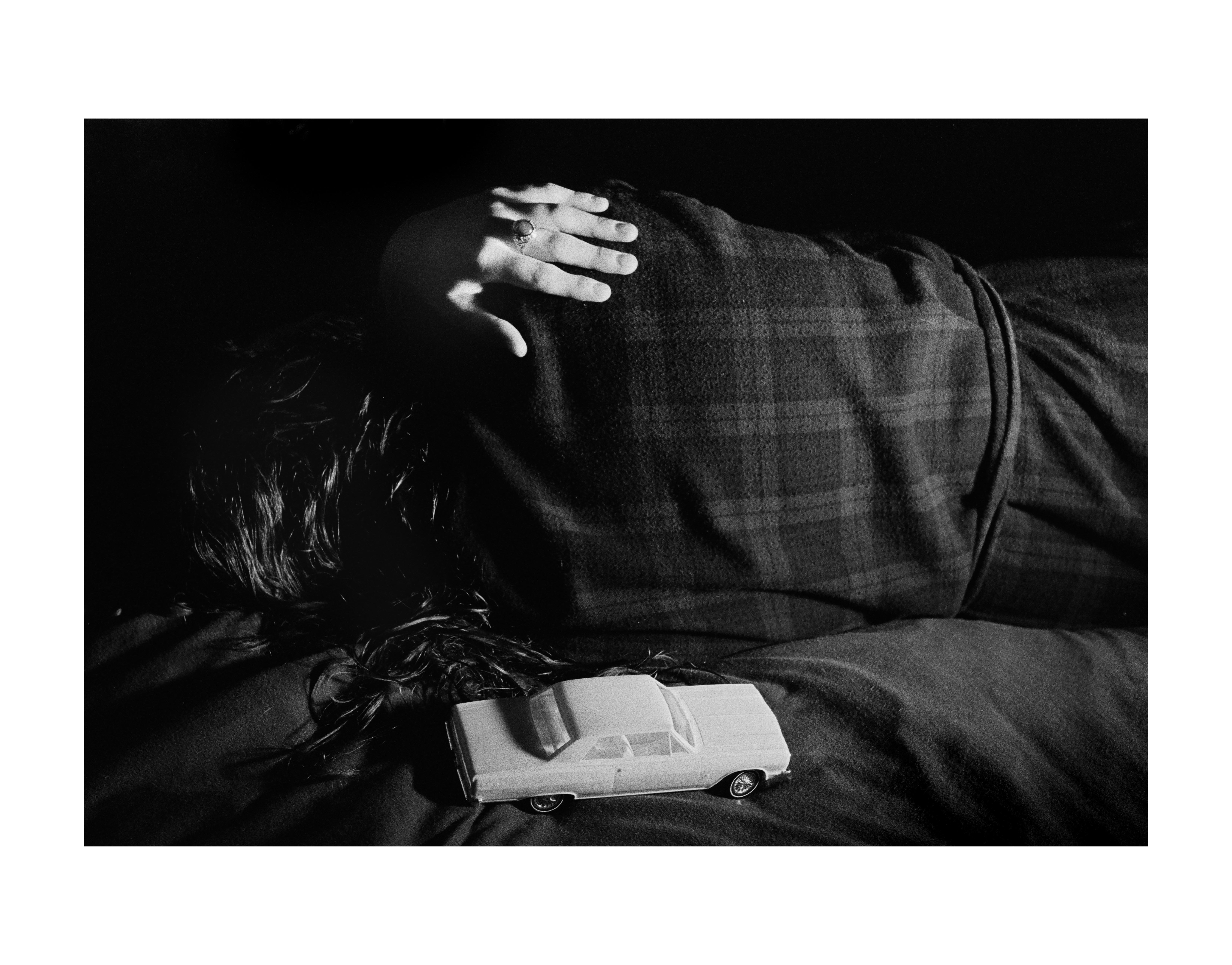 Black and white photo of a person lying on their side, back to the camera in a flannel robe, hand with ring on shoulder, long dark hair touching a toy car in the foreground