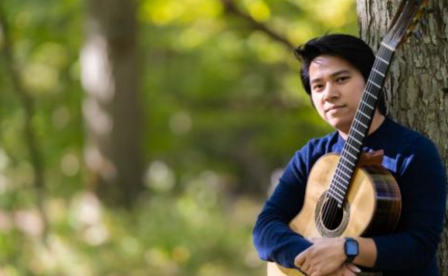 Classical Guitarist An Tran holds guitar and leaning against a tree in a wooded area.