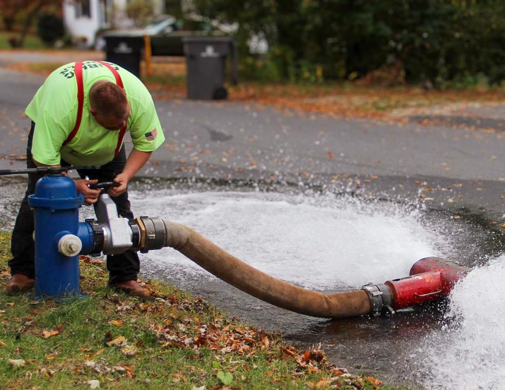 DPW Employee bends over an open hydrant to flush the lines. A hose connected to the hydrant expels water onto the street.