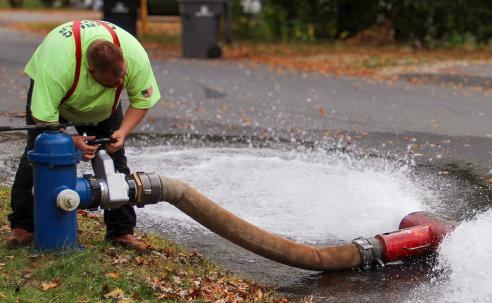 DPW Employee bends over an open hydrant to flush the lines. A hose connected to the hydrant expels water onto the street.